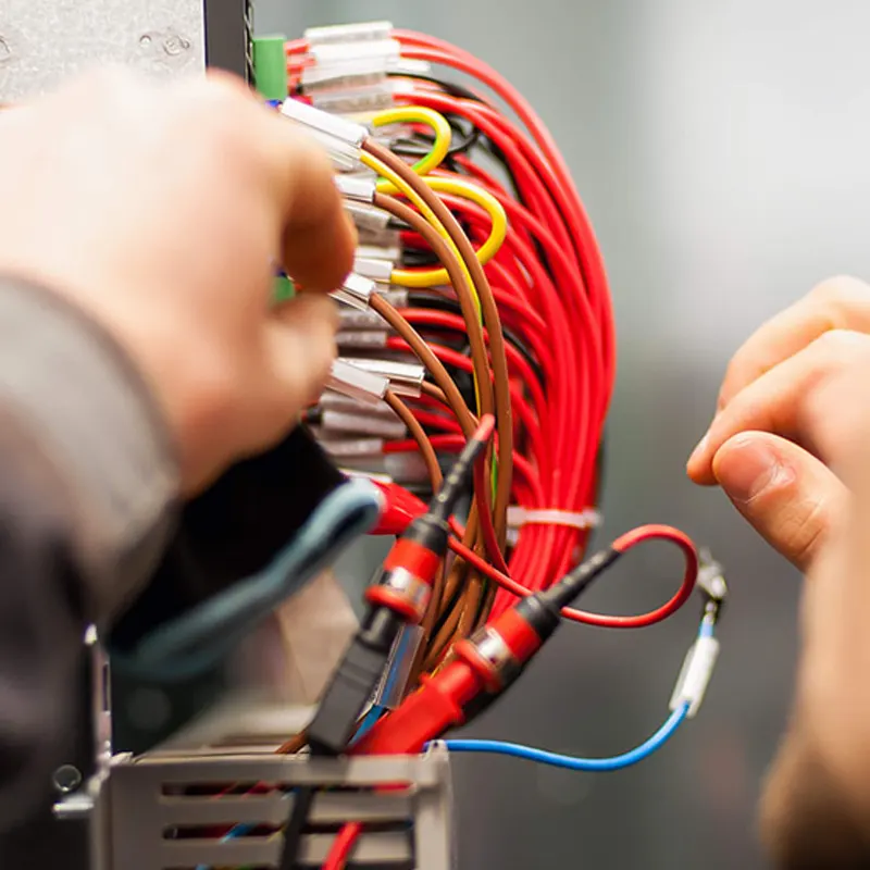 Two hands handle a bundle of red, yellow, and brown wires connected to electronic equipment on the homepage, focusing on the intricate wiring details.